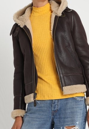 Personne portant une veste en peau retournée marron foncé avec une doublure en fourrure beige, un pull col roulé côtelé jaune et un jean bleu déchiré.