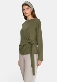 Oliwkowa bluzka z długim rękawem z bocznym wiązaniem. Miękki, lekki materiał, okrągły dekolt i luźny krój. Połączona z jasnobeżowymi spodniami.