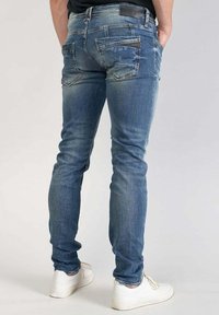 Jeans aderenti blu con una texture sbiadita, caratterizzati da accessori in metallo, due tasche posteriori e un design leggermente usurato. Abbinati a sneaker bianche.