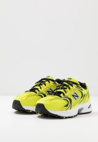 Jasně žluté sportovní boty s černými akcenty a síťovaným svrškem. Mají silné podrážky, odolný vzorek a výrazné boční logo 'N'.