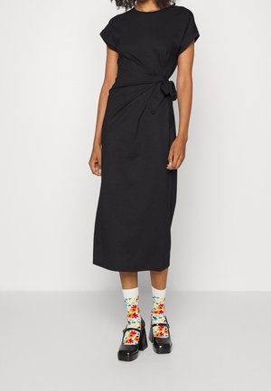 Kobieta w czarnej sukience midi z wiązaniem z boku, kolorowe kwieciste skarpetki i czarne botki na platformie Mary Jane, stojąca na jednolitym tle.