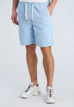 Mężczyzna ubrany w jasnoniebieskie spodenki z troczkiem i białe buty sportowe, stojący z rękami w kieszeniach na jednolitym tle.