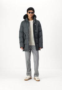 Veste en duvet gris foncé, avec une capuche doublée en fausse fourrure, une poche poitrine zippée et des fermetures à pression. Associée à un pantalon gris clair et des baskets blanches.