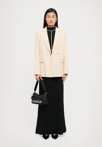 Woman stands facing forward wearing a cream blazer over a black high-neck top and long black skirt, holding a black Love Moschino handbag.