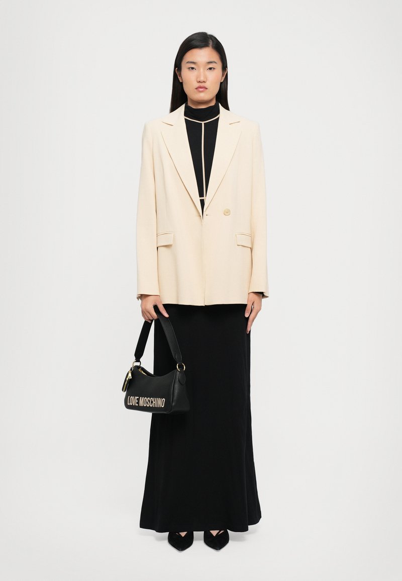 Woman stands facing forward wearing a cream blazer over a black high-neck top and long black skirt, holding a black Love Moschino handbag.