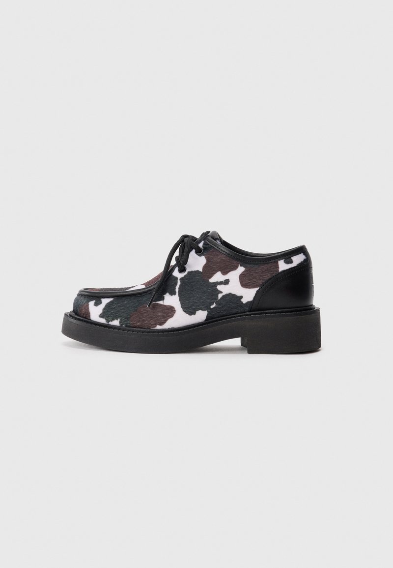 Chaussures en cuir avec un imprimé vache noir, blanc et brun, dotées d'une semelle en caoutchouc noir épaisse et d'accents noirs au niveau du talon et des lacets.