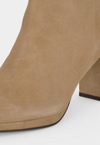 Gros plan d'une bottine en daim beige avec un bout arrondi et un talon épais de hauteur moyenne sur fond blanc.