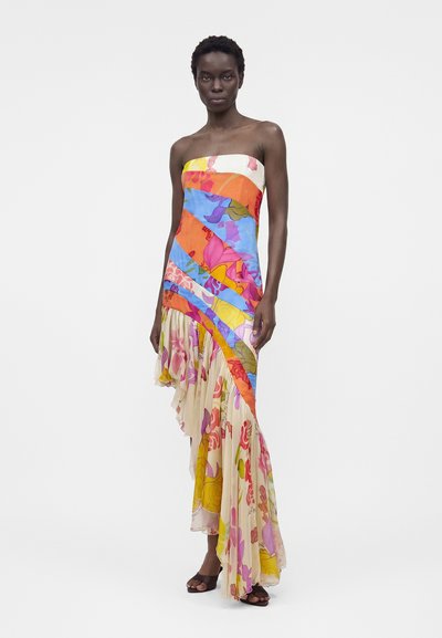 Μοντέλο που φοράει πολύχρωμο μακρύ φόρεμα χωρίς τιράντες με ασύμμετρο βολάν στο τελείωμα και σκούρα πέδιλα με ανοιχτά δάχτυλα σε μονόχρωμο φόντο.