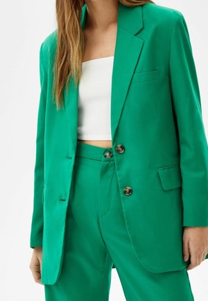 Femme portant un costume vert vif avec un blazer boutonné et un pantalon assorti, associé à un crop top blanc côtelé.
