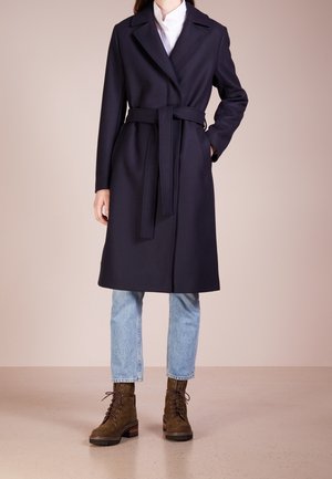 Person iført en knælængde mørk marineblå bæltefrakke, lyseblå jeans og brune snørestøvler ved anklerne, stående mod en beige baggrund.