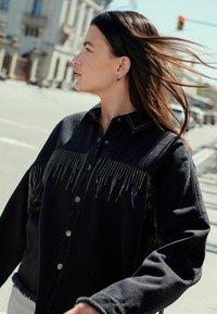 Juodas džinsinis švarkas su fringing akcentais, mygtukais snap ir apykakle. Audinys yra lygus, su metaliniais akcentais priekyje.