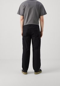 Donkergrijze T-shirt gecombineerd met losse zwarte cargobroek met zijzakken, elastische tailleband en verstelbare boord. Zwarte schoenen zichtbaar.