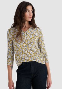 Top floreale a scollo a V in senape con motivi di fiori neri e bianchi. Caratterizzato da maniche lunghe e una vestibilità comoda. Abbinato a jeans in denim scuro.