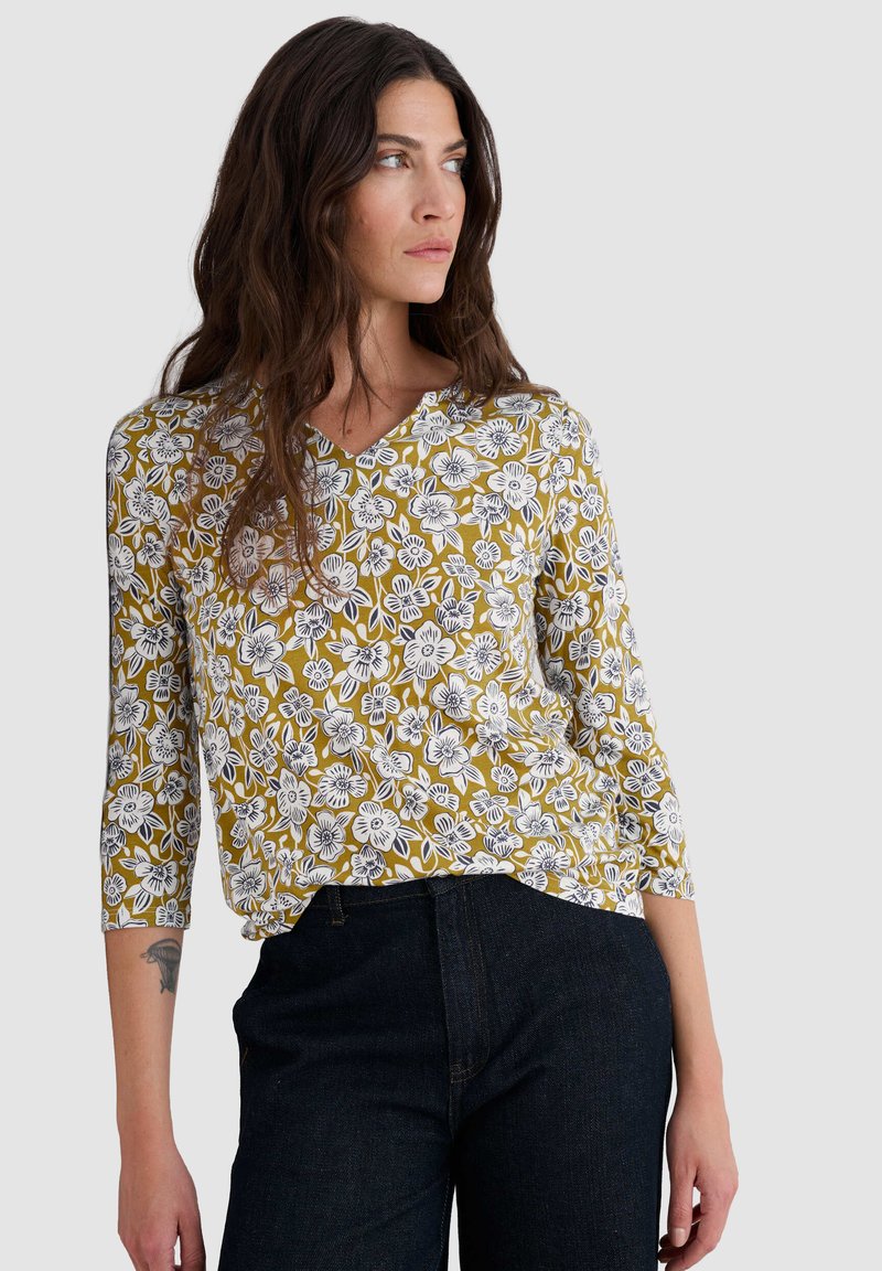 Top floreale a scollo a V in senape con motivi di fiori neri e bianchi. Caratterizzato da maniche lunghe e una vestibilità comoda. Abbinato a jeans in denim scuro.