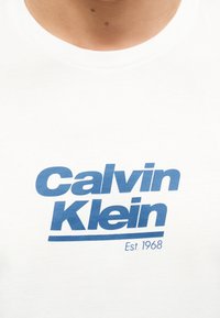 Λευκό T-shirt με μπλε λογότυπο "Calvin Klein" και κείμενο "Est. 1968" στο στήθος, φορεμένο από ένα άτομο με ορατό λαιμό και ώμους.