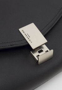 Černá kožená taška s hladkým povrchem, opatřená obdélníkovým stříbrným kovovým zapínáním a prošitým okrajem. Název značky je vyražen na zapínání.