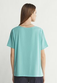 Turkos kortärmad topp med lös passform, rund halsringning och slät yta, tillsammans med ett guldhalsband i nacken.