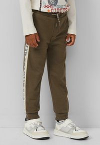 Braune Sweatpants mit weißen Seitenstreifen und bedrucktem Text. Hergestellt aus weichem Stoff, haben sie ein tailliertes Design und elastische Bündchen. Kombiniert mit weißen und grauen Sneakers.