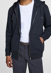 Sudadera con capucha y cremallera en azul marino oscuro. Cuenta con dos bolsillos frontales, puños de punto y una textura suave. Llevada sobre una camiseta blanca.