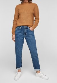 Roodbruine geribbelde gebreide trui gecombineerd met lichtblauwe denim jeans met opgerolde mouwen en witte sneakers. De look is casual en ontspannen.