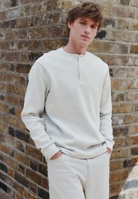 Pitkähihainen, vaaleanharmaa henley-paita teksturoidulla kuviolla, jossa on kolme nappia kaula-aukossa, yhdistetty vaaleisiin housuihin.