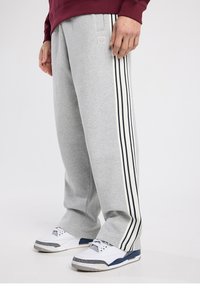 Un pantalon de survêtement gris avec une taille élastique, des bandes latérales bleu marine et crème, et un petit logo à l'avant. Porté avec des chaussures de sport blanches.