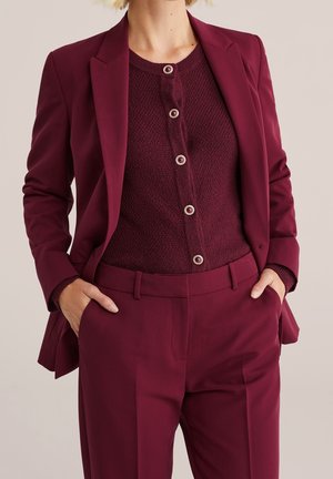 Woman wearing a burgundy tailored blazer, matching high-waist pants, and a textured buttoned cardigan with metallic buttons. Hands in pockets.