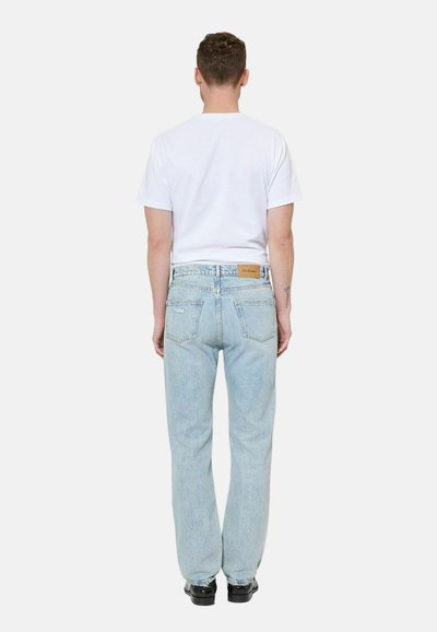 Homme debout de dos, portant un t-shirt blanc uni à manches courtes, un jean bleu clair avec un patch de marque en cuir, et des chaussures noires.