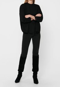 Blouse noire à manches longues avec un col haut et des détails froncés, associée à un jean en denim noir et des bottes hautes en daim noir.