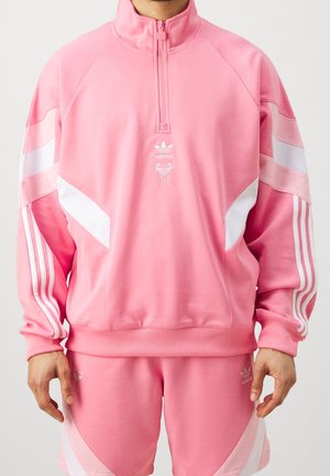 Osoba ubrana w różowy dres Adidas z białymi paskami i pulower z suwakiem do połowy, stojąca na tle jednolitego jasnego tła.