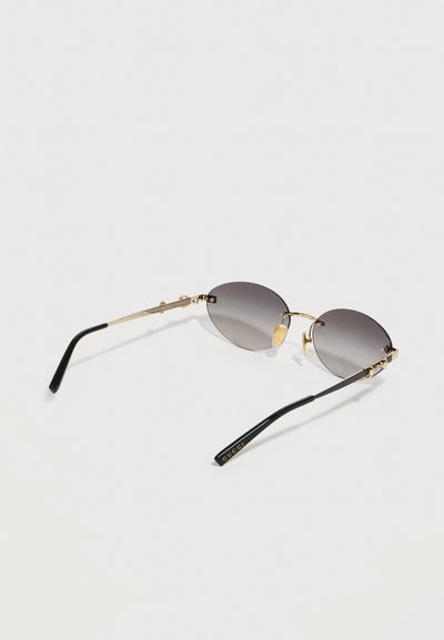 Οβάλ γυαλιά ηλίου χωρίς πλαίσιο με γκρι φακούς με διαβάθμιση, χρυσή μεταλλική γέφυρα και βραχίονες, και μαύρα ακουστικά με χρυσλό λογότυπο Gucci.