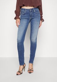 Jeans de mezclilla azul ajustados con una cintura de altura media, un desgaste sutil y estilo de cinco bolsillos, combinados con unos zapatos de tacón alto negros con tiras.