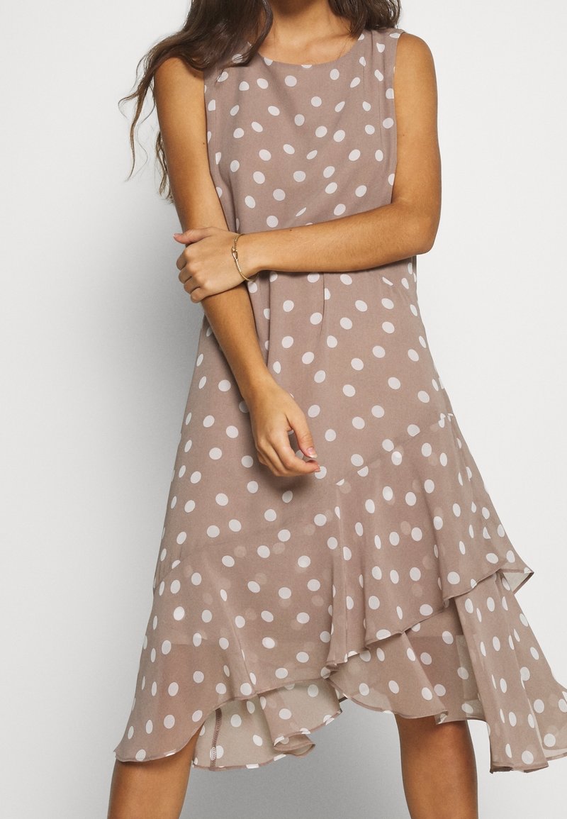 Braunes ärmelloses Kleid mit weißen Punkten, geschichteter Rüschensaum, aus einem leichten Stoff. Arme des Models verschränkt, ein Armband am Handgelenk.