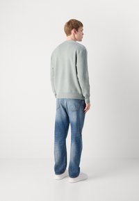 Helle grüne, gerippte Strickjacke mit langen Ärmeln, kombiniert mit hellblauen Jeans, getragen mit weißen Sneakers. Sicht von hinten.