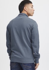 Grå hettegenser med langermet og ribbestrikkede mansjetter, som viser en jevn tekstur, kombinert med mørkejeans. Synlige sømdetaljer ved kanten.