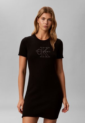 Kobieta z długimi falującymi włosami, ubrana w krótką czarną sukienkę z logo „Calvin Klein” na piersi, stojąca na tle jednolitego szarego tła.