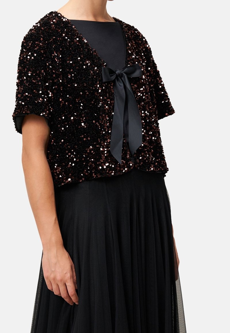 Femme portant un top noir à sequins à manches courtes avec un nœud papillon en satin sur une robe noire avec une jupe transparente et plissée.