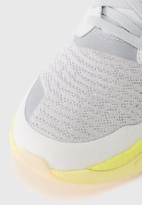 Detalhe em close-up de um bico de sapatilha de malha branca, com sobreposições de camurça cinza claro e detalhe de sola amarelo pálido.