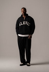 Schwarzer Fleece-Pullover mit weißem "BLKVIS"-Stick, ausgestattet mit einem Reißverschlusskragen. Kombiniert mit schwarzen maßgeschneiderten Hosen und schwarz-weißen Schuhen.
