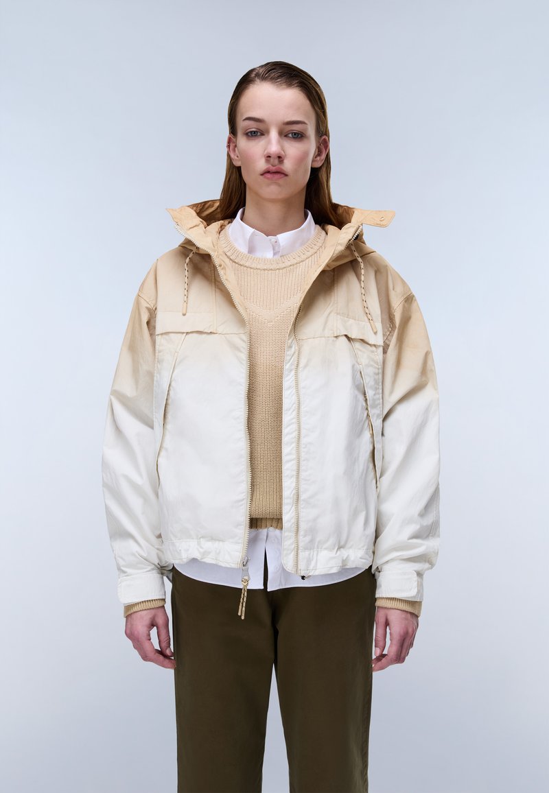 Ung kvinde iført en beige og cremefarvet hættejakke over en tanfarvet sweater og hvid skjorte, kombineret med mørke olivengrønne bukser, stående mod en ensartet baggrund.