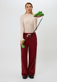 Modell som holder en grønn roterende telefon, iført en tettsittende, teksturert, kremfarget langermet topp og burdeos høyt midje joggebukser med sideaksenter.