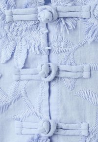 Tissu bleu clair avec broderie florale et trois boutons nœuds en tissu disposés verticalement, montrant une texture et une couture détaillées.