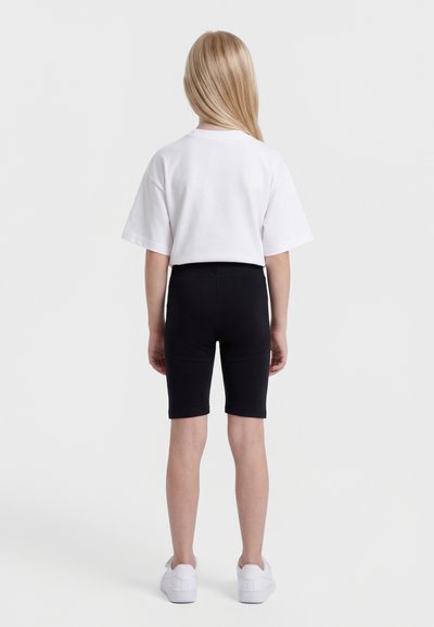 Παιδί με μακριά ξανθά μαλλιά, που φοράει λευκό κοντομάνικο μπλουζάκι, μαύρο σορτς μέχρι το γόνατο και άσπρα αθλητικά παπούτσια, όρθιο με την πλάτη στραμμένη προς τα εμπρός.
