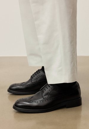 Čierne kožené brogue topánky nosené s bielymi nohavicami na leštenej podlahe, zdôrazňujúce klasické detaily formálnej obuvi.