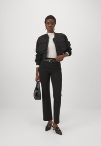 Schwarze, cropped Bomberjacke mit doppelten Fronttaschen, weißes Oberteil, hochtaillierte schwarze Hose und schwarze Slingback-Schuhe. Hält eine schwarze Handtasche.