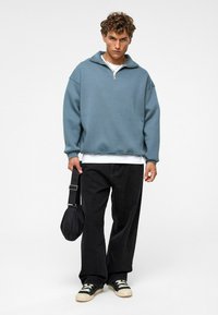 Lichtblauwe sweater met een ritskraag, gedragen over een wit shirt, gecombineerd met zwarte jeans en zwarte schoenen, en een zwarte tas vasthoudend.