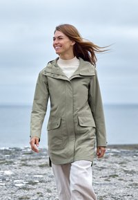 Kaki regnjacka med huva, framsida dragkedja och tryckknappsfickor. Slät textur, mittlånga design med en avslappnad passform över ljusfärgade byxor.