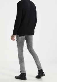 Man med svart stickad tröja, grå slim-fit jeans och svarta läderanklar boots som går bort från en enfärgad vit bakgrund.