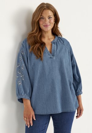 Kvinde med langt rødt hår iført en løs denimbluse med blomsterbroderi på den ene ærme og mørkeblå jeans, smilende blidt.