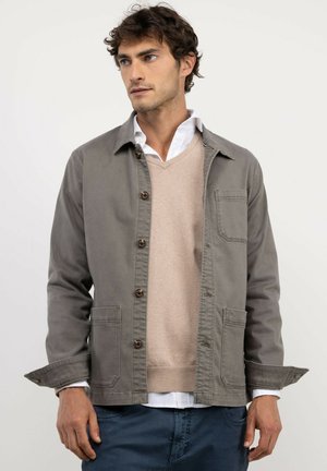 Joven con una chaqueta de camisa gris con botones sobre un suéter beige y una camisa blanca, de pie con expresión neutra, mirando hacia la izquierda.